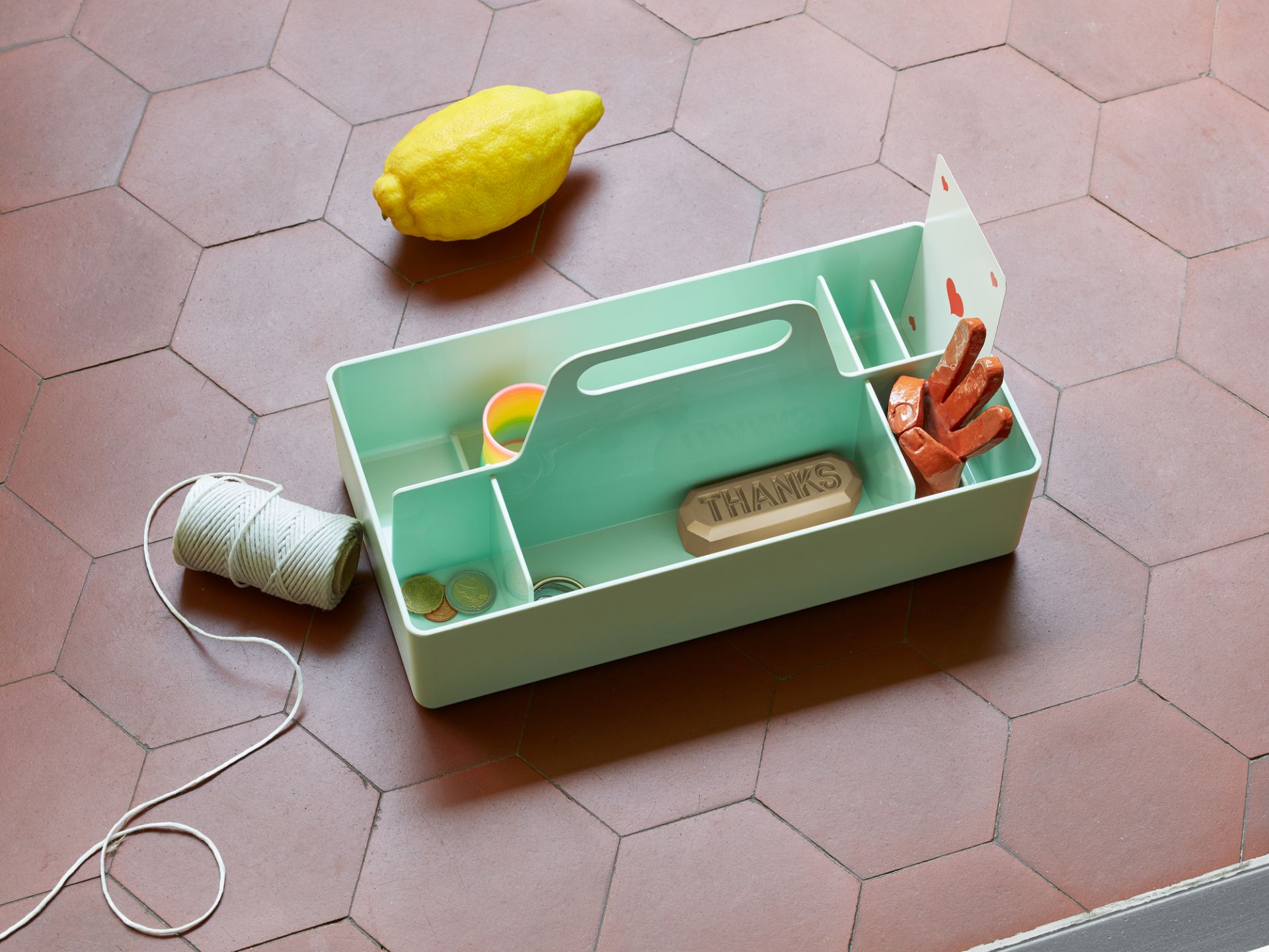 Helle Vitra Toolbox Organisationsbox, gefüllt mit Büroartikeln, auf einem Terrakotta-Fliesenboden mit Zitrone und Garn.