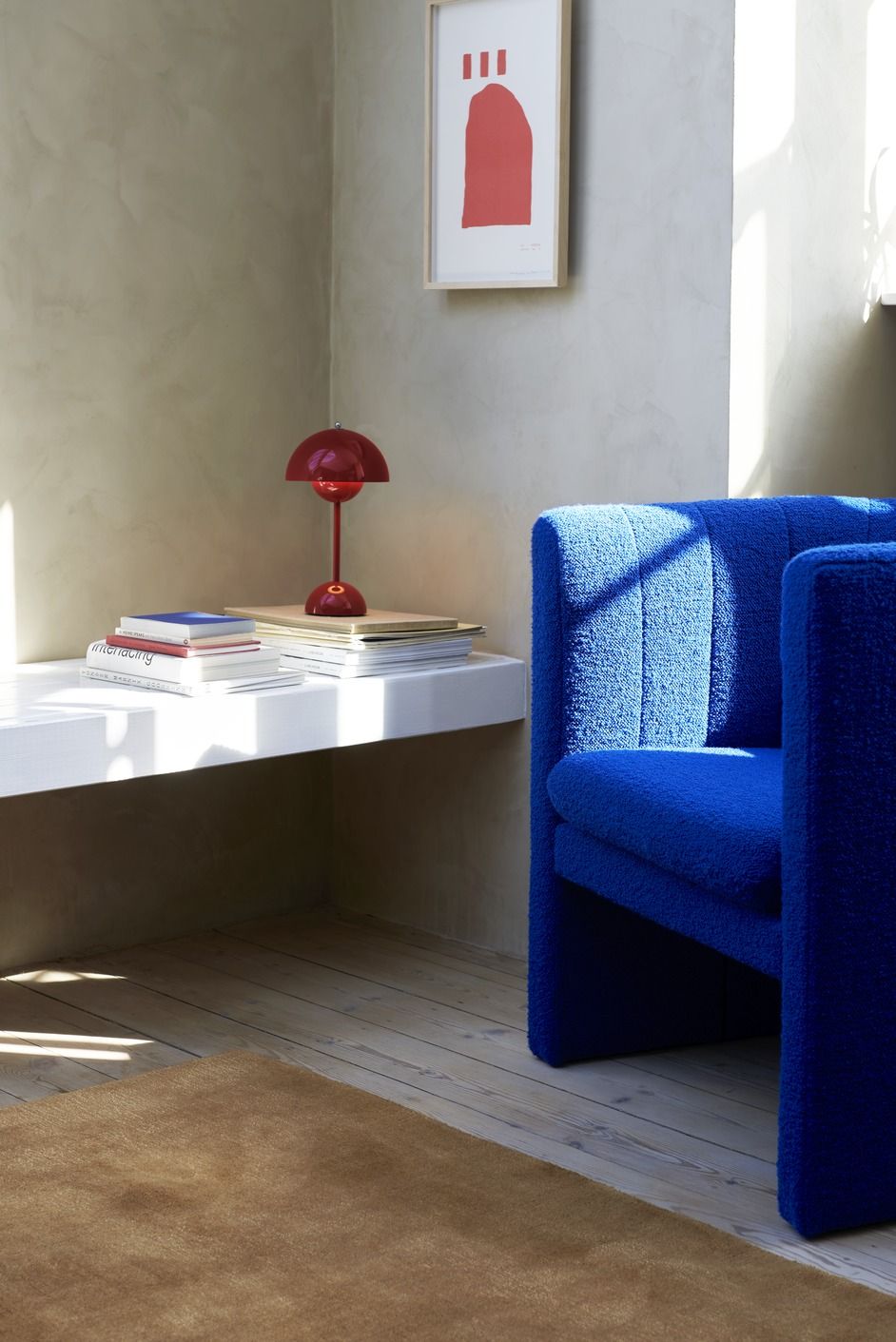 Flowerpot VP9 Tischleuchte in Vermilion Red auf Bücherstapel neben blauem Sessel.