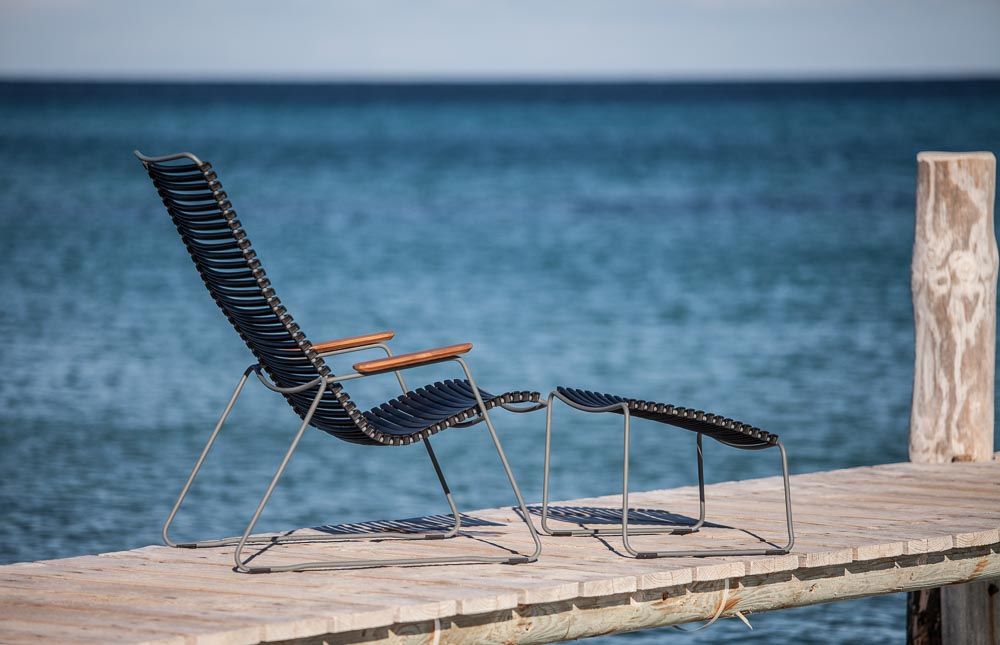 Dunkelgrauer Click Lounge Chair Sessel von Houe mit Fußhocker auf Holzsteg vor blauem Meer.