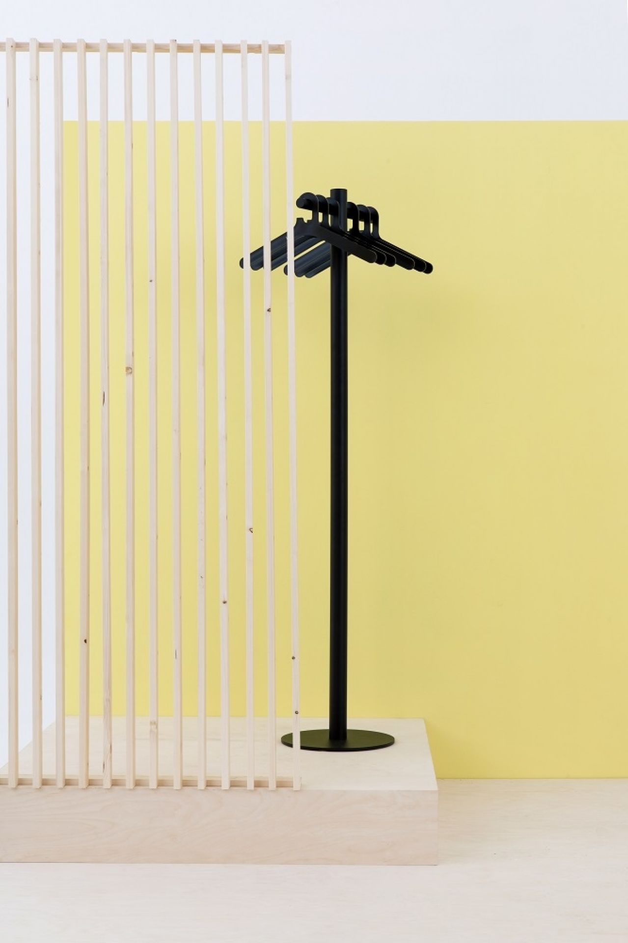 Schwarzer Cascando Pole Garderobenständer mit Kleiderbügeln vor gelbem Hintergrund und Holzpaneel. Moderner Garderobenständer für Büro und Zuhause.