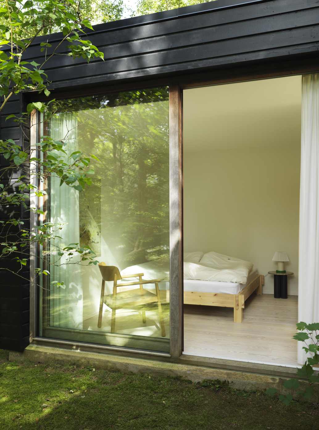 Ansicht durch ein Fenster auf ein Schlafzimmer mit einem Normann Copenhagen Beistelltisch neben einem Bett.