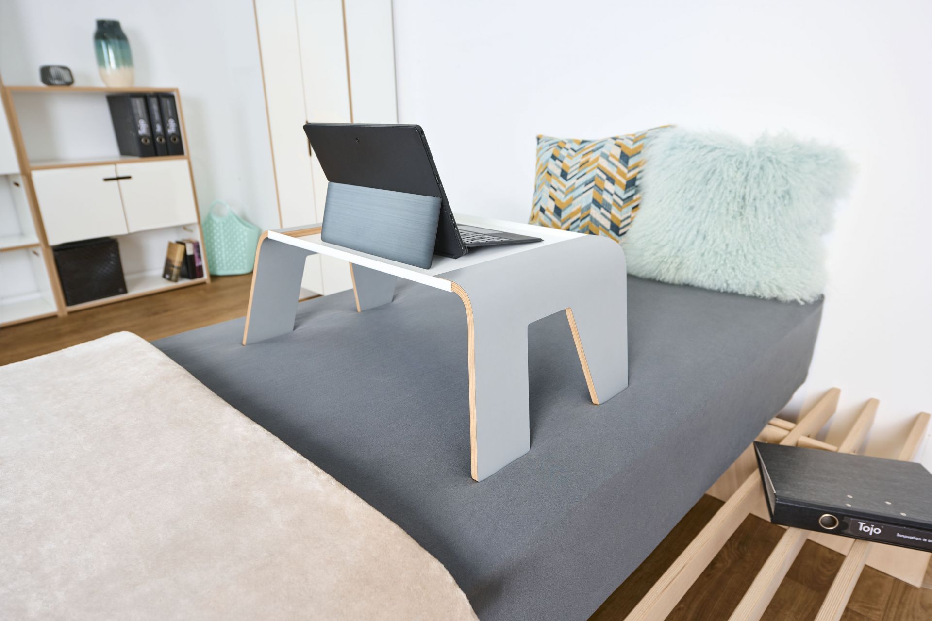 Tojo Notebook-Tablett auf Bett mit Laptop, Kissen und grauem Bettlaken für entspanntes Arbeiten.