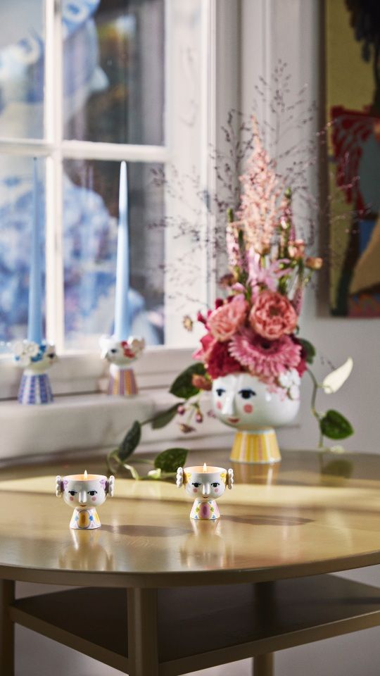 Zwei Bjorn Wiinblad Eva Teelichthalter im 2er-Set auf Tisch, Kerzenlicht und Blumenvase im Hintergrund.