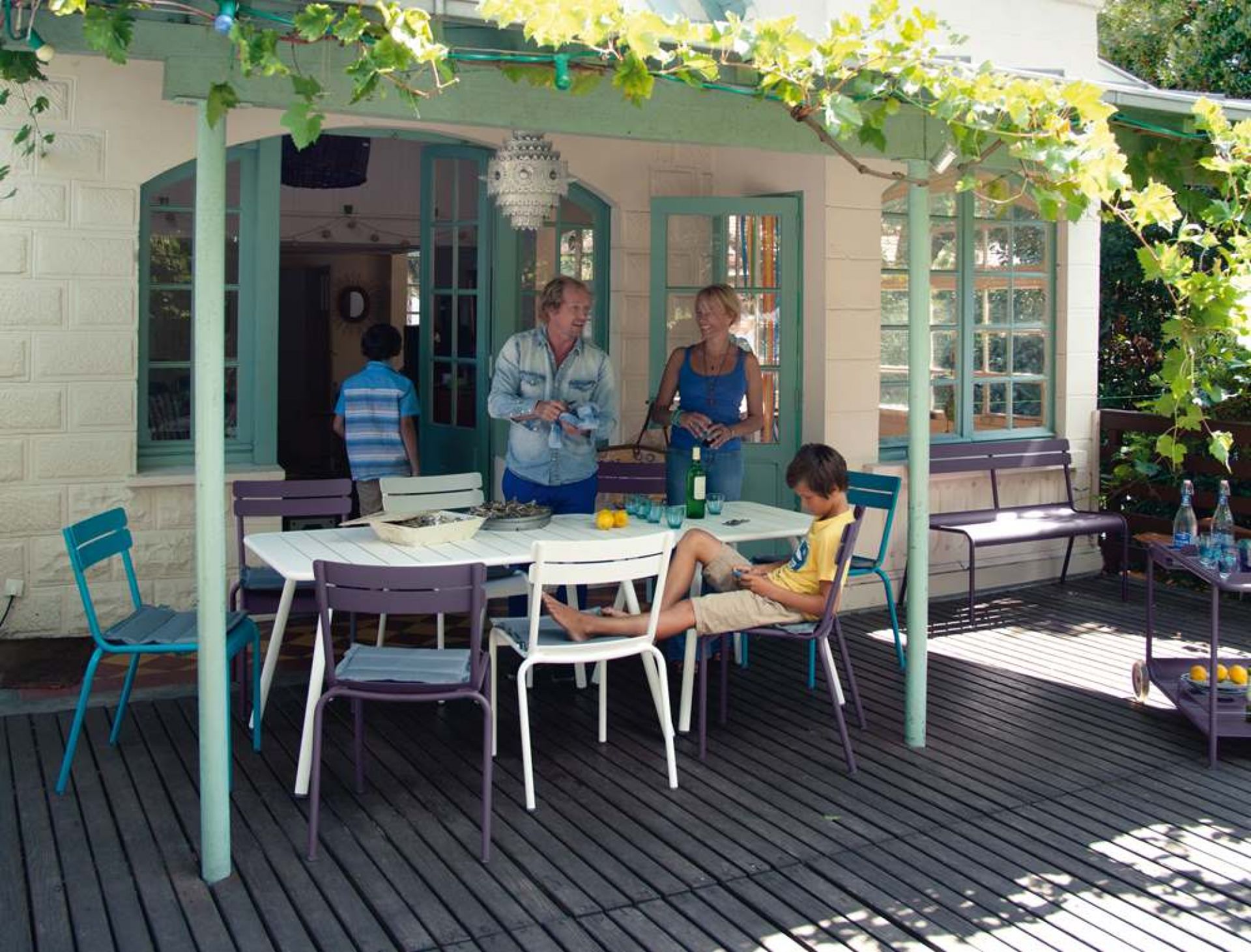 Fermob Luxembourg Gartenmöbel-Set: Tisch, Stühle, Bank und Servierwagen auf einer Terrasse mit Familie.