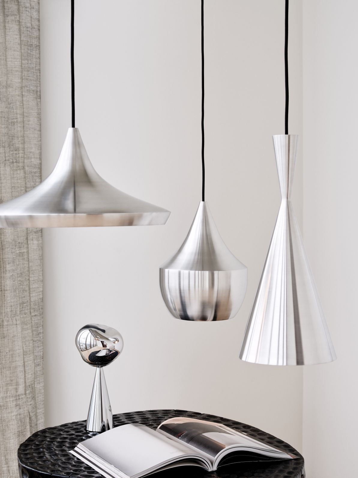 Drei moderne Tom Dixon Pendelleuchten aus gebürstetem Aluminium hängen über einem Tisch mit Lampe.