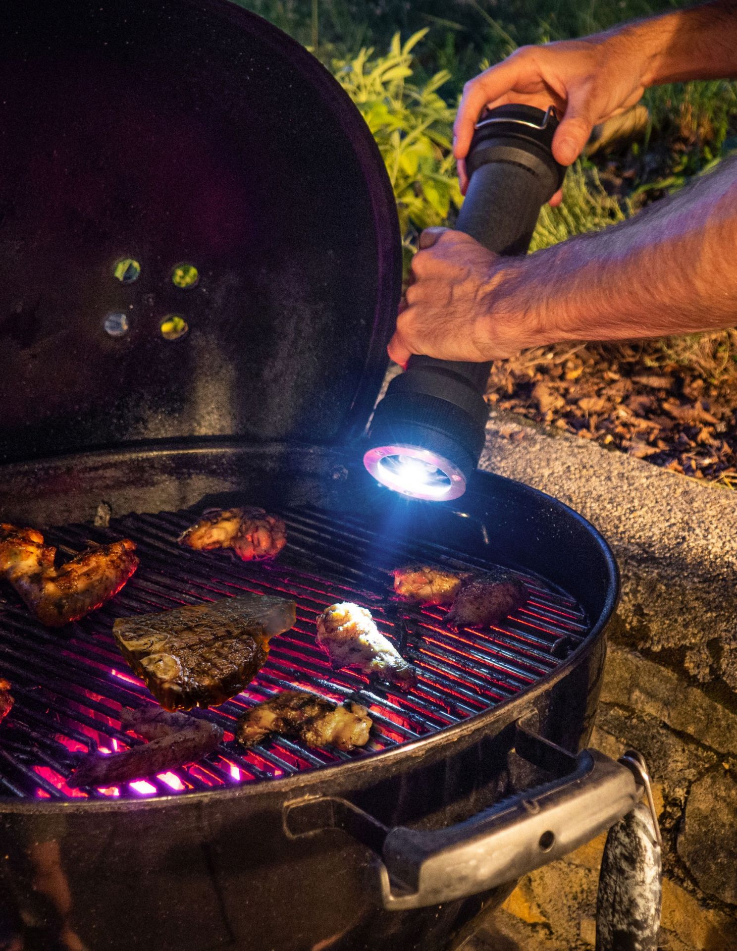 Mann grillt Steaks und Hähnchen auf einem Holzkohlegrill und leuchtet mit einer Taschenlampe.