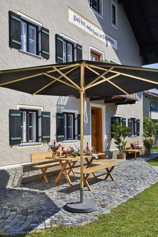 Weishäupl Klassiker Sonnenschirm mit schwarzem Schirmbezug und Holzgestell vor einem traditionellen Haus mit Holzbänken.