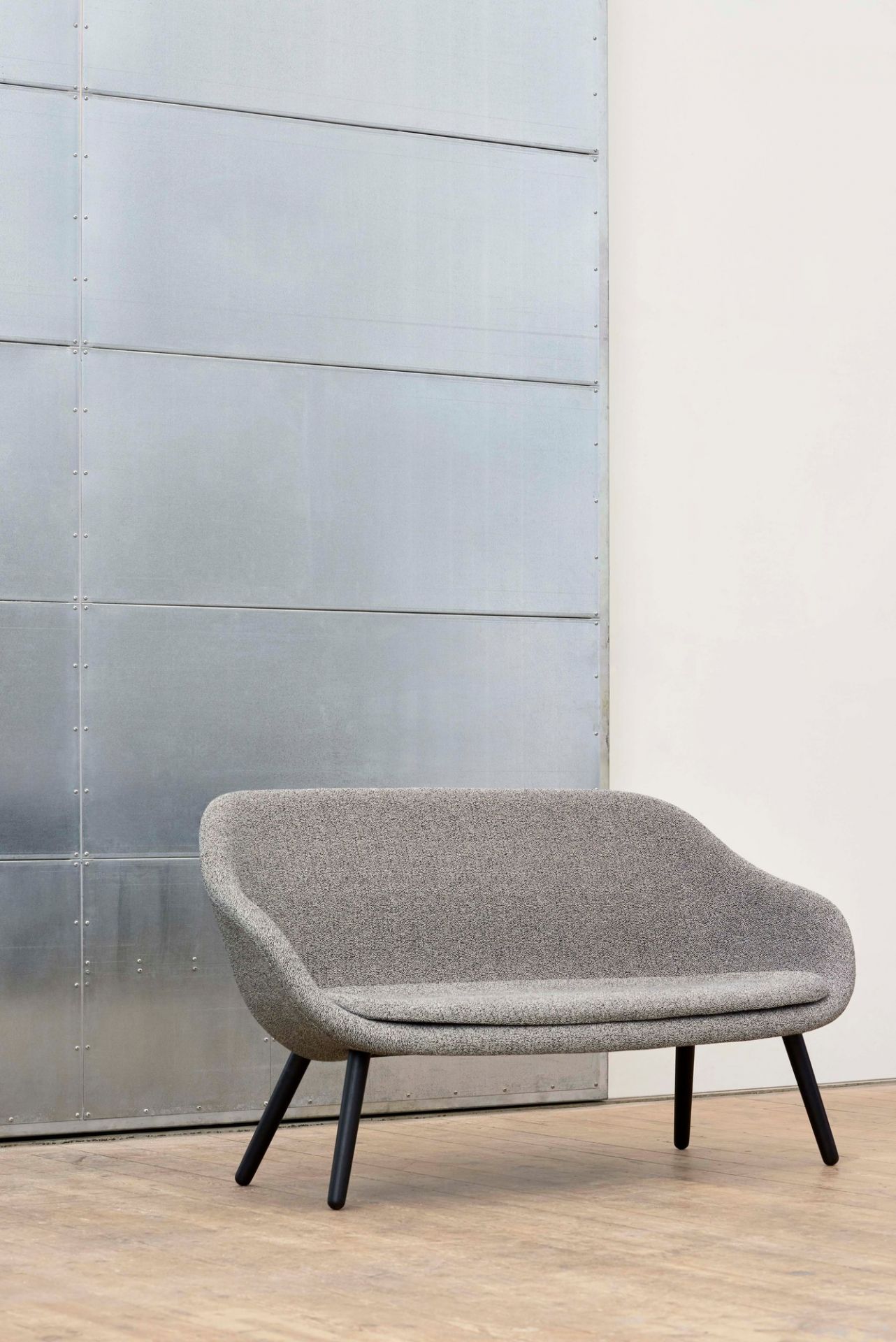 Graues Hay About a Lounge Sofa AAL mit schwarzen Holzbeinen vor Metallwand.