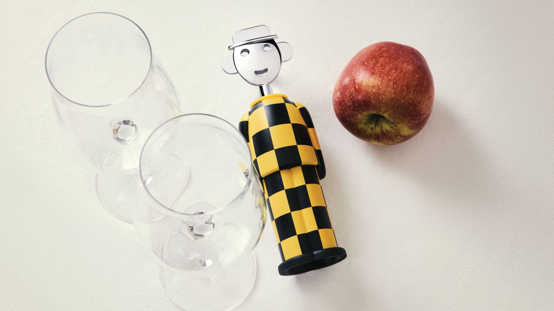 Alessi Alessandro M. Korkenzieher, gelb-schwarz kariert, mit Apfel und Weingläsern auf weißem Hintergrund.