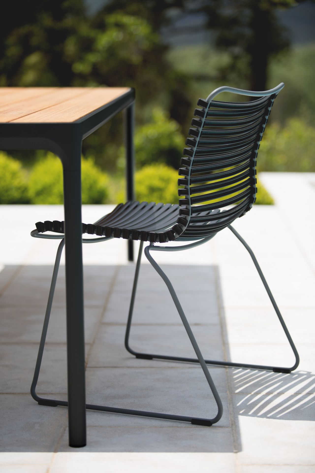 Houe Click Dining Stuhl ohne Armlehne, graues Outdoor-Design mit Lamellen, neben einem Gartentisch.
