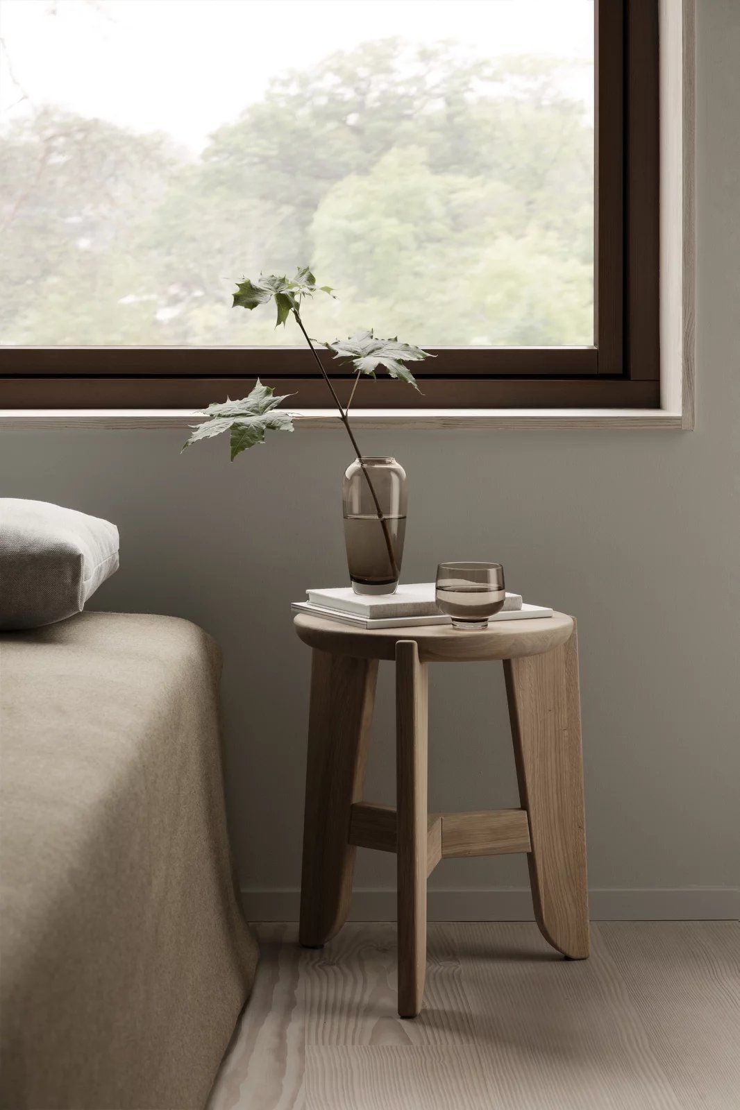 Eli Stool Sitzhocker von Blomus aus hellem Holz mit Deko-Elementen neben einem Bett.