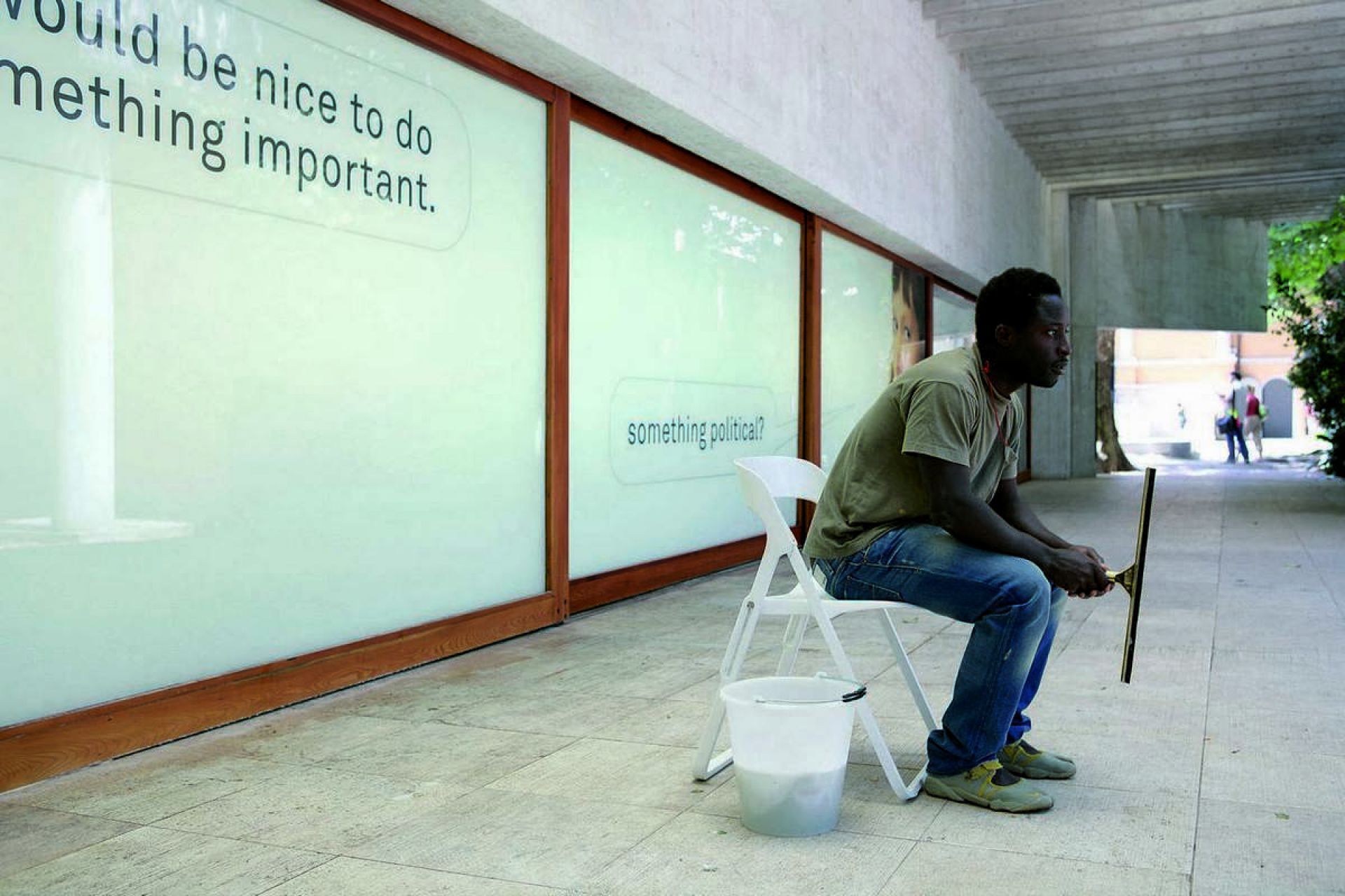 Mann sitzt auf weißem Klappstuhl mit Eimer und Fensterabzieher, Fenster mit Text im Hintergrund.
