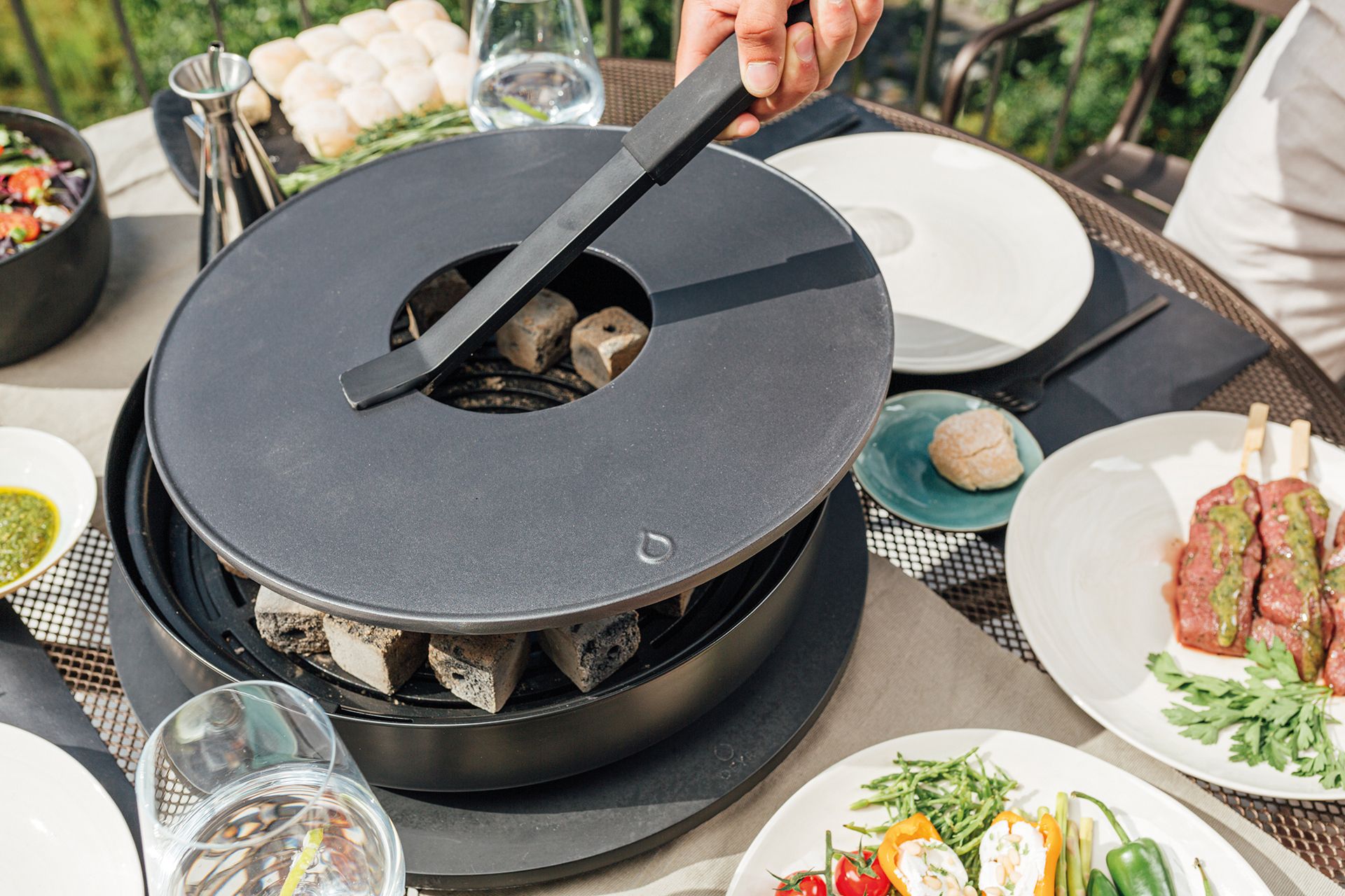 Tabl´O Tischgrill von Ofyr mit Grillzange und Holzkohle, umgeben von Speisen auf dem Tisch.