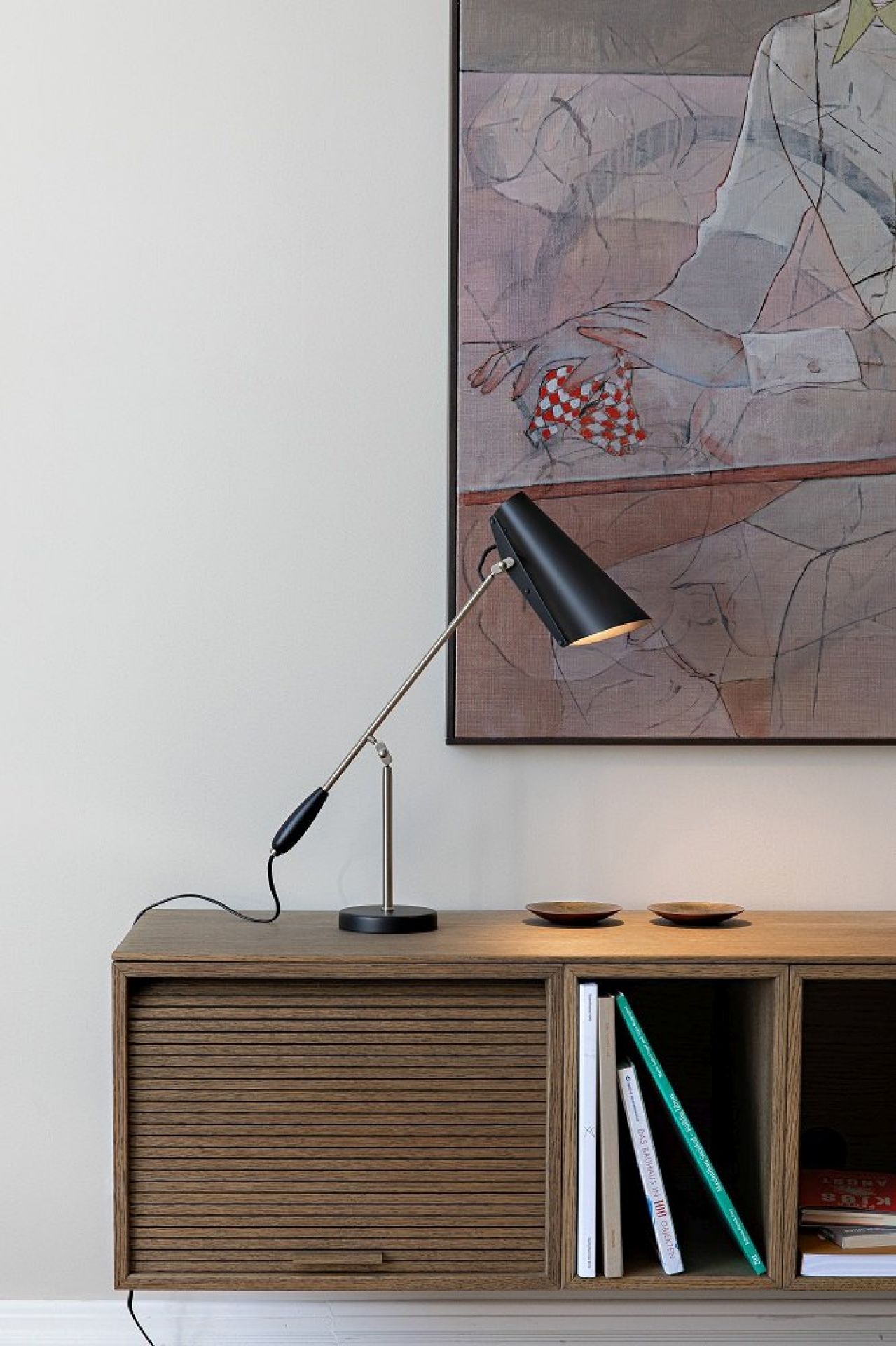 Schwarze Birdy Tischleuchte von Northern auf einem Holzschrank mit Kunstwerk im Hintergrund. Design Lampe für Wohnzimmer.