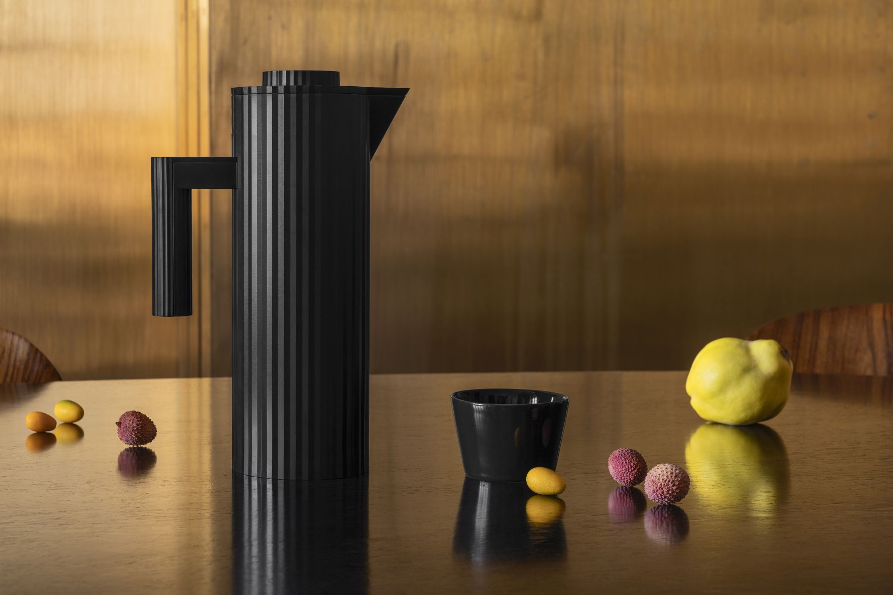 Graue Alessi Plissé Isolierkanne MDL12 mit gerippter Oberfläche, Tasse und Früchten auf dunklem Holztisch.