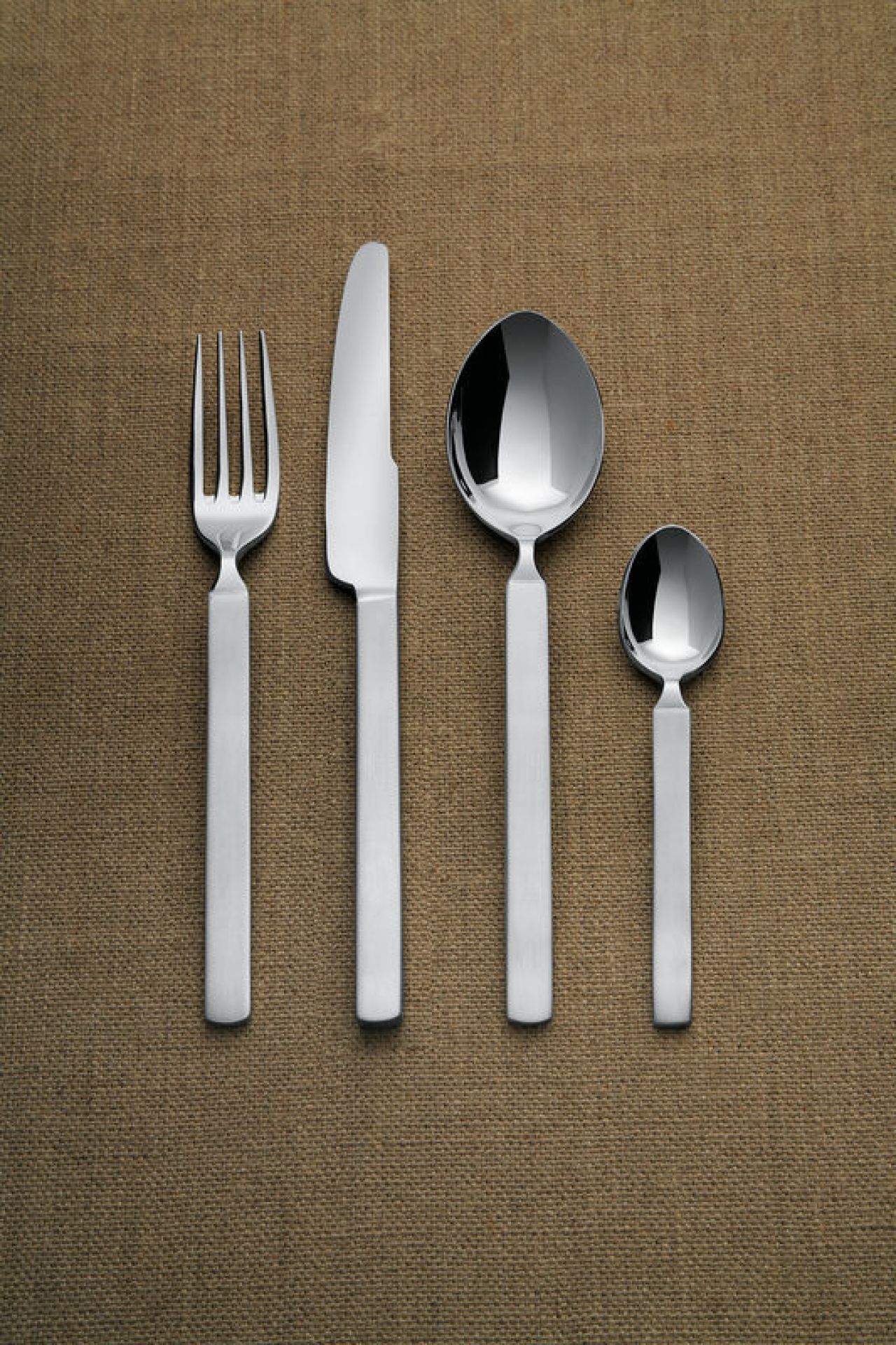 Alessi Dry Besteckset: Messer, Gabel, Esslöffel und Teelöffel aus Edelstahl auf braunem Stoffhintergrund.
