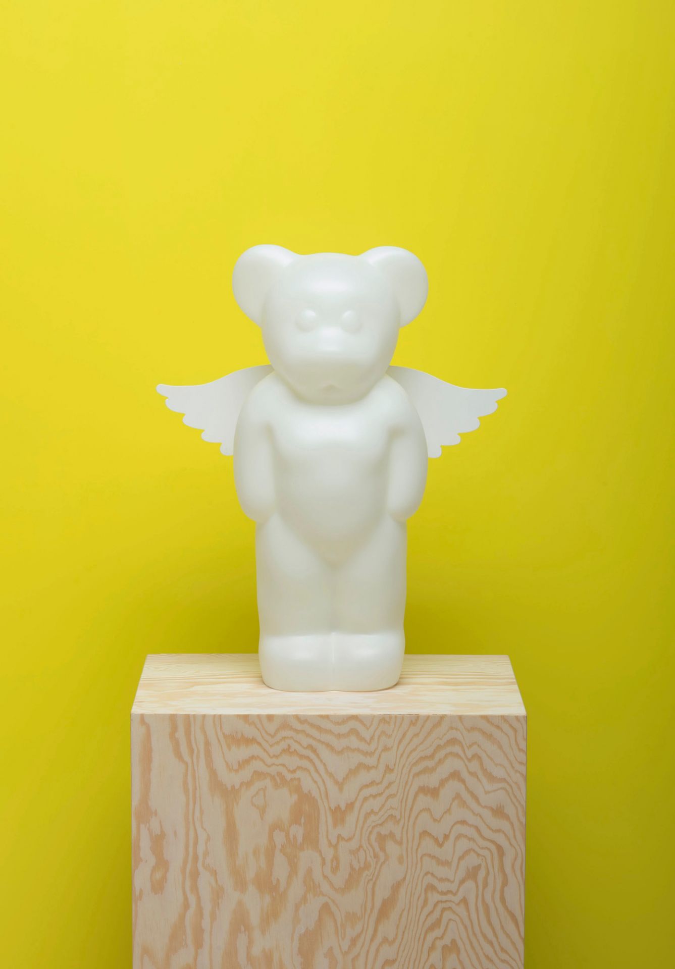 Lumibär Stehleuchte mit weißen Engelflügeln auf einem Holzsockel vor gelbem Hintergrund.