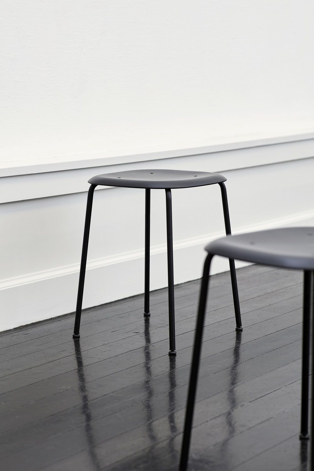 Schwarzer Hay Soft Edge Hocker mit drei Beinen auf dunklem Holzboden vor weißer Wand.