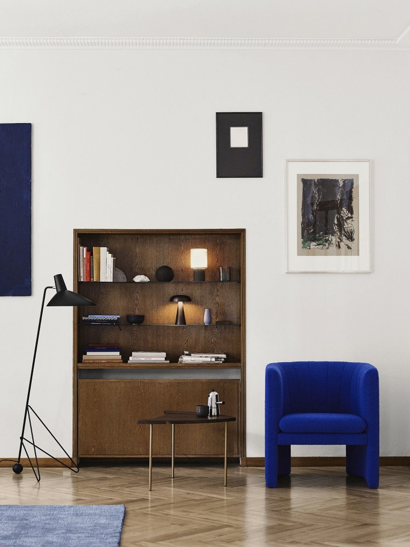 AndTradition Manhattan SC52 Akkuleuchte auf Regal, neben Stehlampe und blauem Sessel in modernem Wohnzimmer.