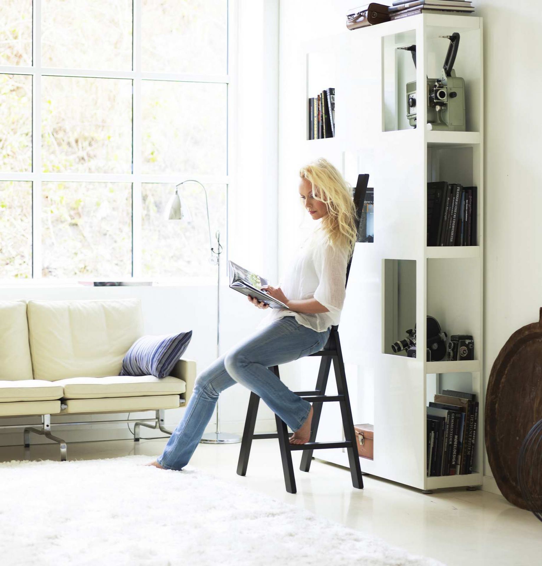Frau liest auf einer schwarzen Hindevadgaard Trittleiter in einem hellen Wohnzimmer mit Sofa und Bücherregal.