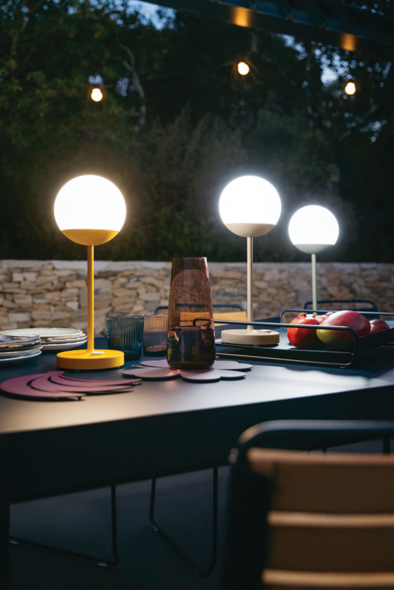 Mooon Tischleuchte von Fermob, anthrazitfarbener Tisch mit Lampen, Gläsern und Obst im Garten.