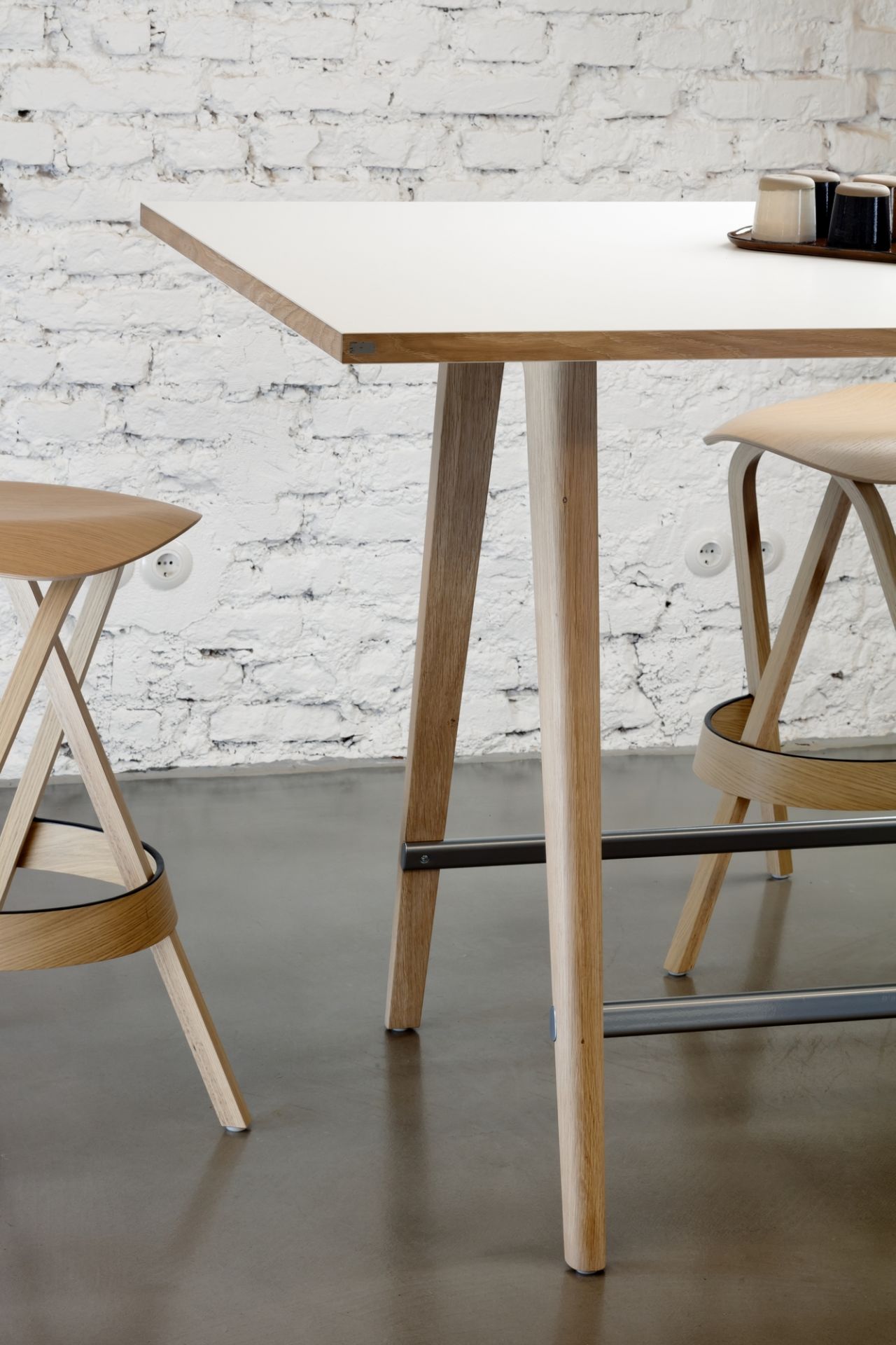 Detailaufnahme des Thonet 1510 Stehtisches mit Barhockern, hellem Holzgestell und weißer Tischplatte vor einer Backsteinwand.