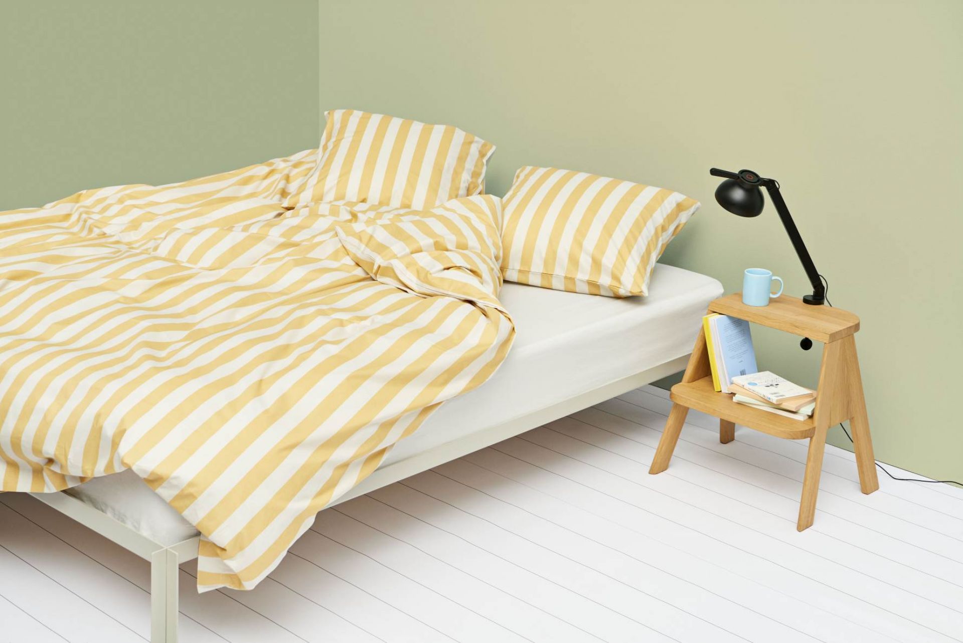 Heller Raum mit Bett und gelb-weiß gestreifter Bettwäsche, daneben ein Hay Butler Hocker als Nachttisch.