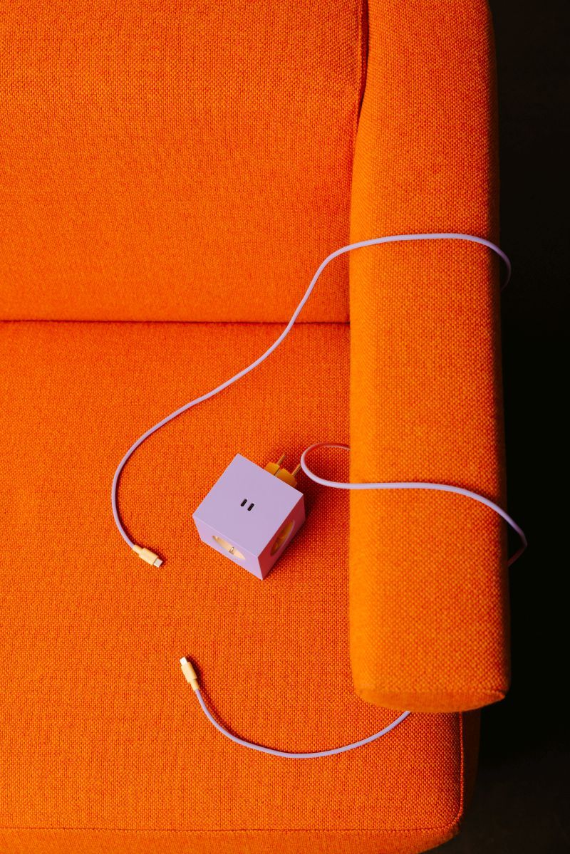 Lila Avolt Square 2 Steckdosenwürfel auf einem orangefarbenen Sessel mit USB-C Kabel.