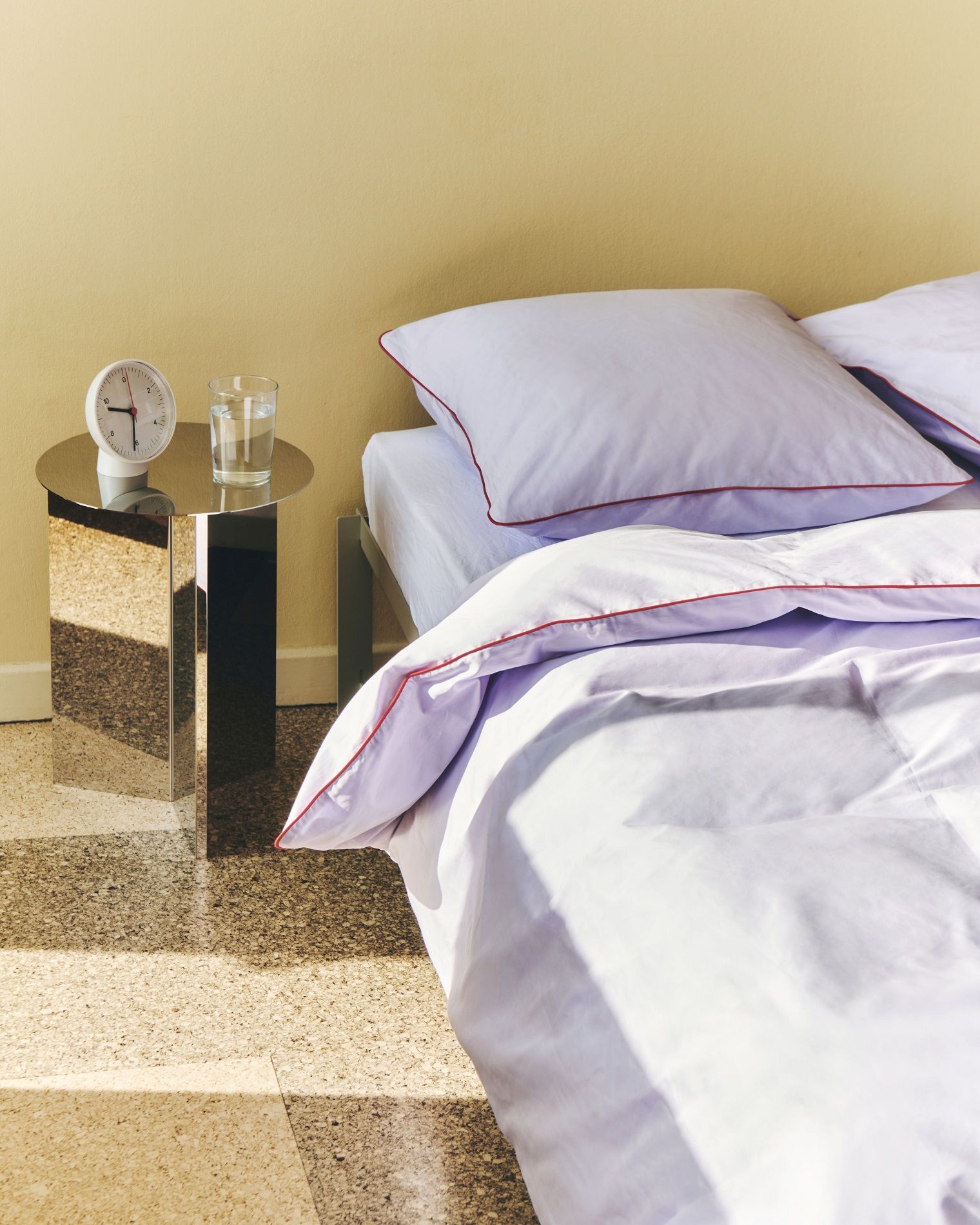 Silberner Hay Slit Table Round High Beistelltisch neben einem Bett mit Uhr und Wasserglas.
