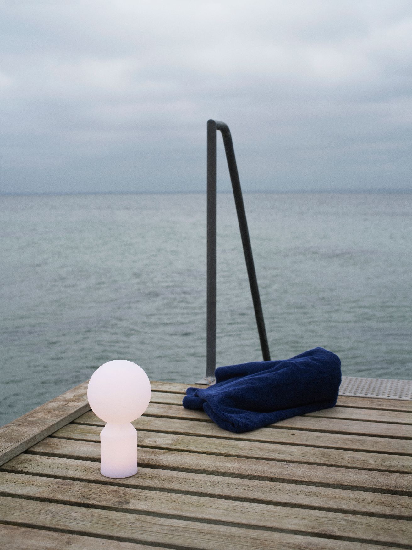 Normann Copenhagen Yo Lamp Small Akkuleuchte auf Holzsteg am Meer, neben blauem Handtuch.